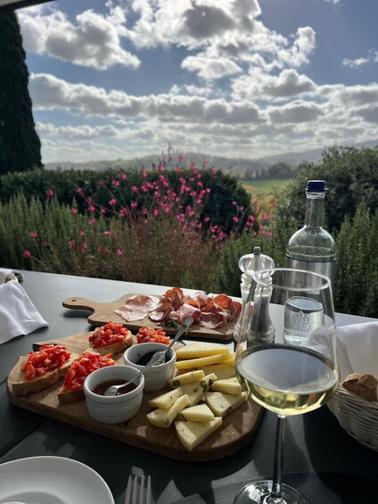A picnic table with charcuterie, cheese, and a glass of white wine, overlooking a scenic landscape with pink flowers and a clear sky. 5