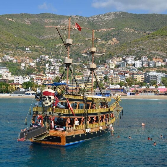 A pirate-themed boat with a skull figurehead, sailing near a coastal town with buildings and hills in the background. 1