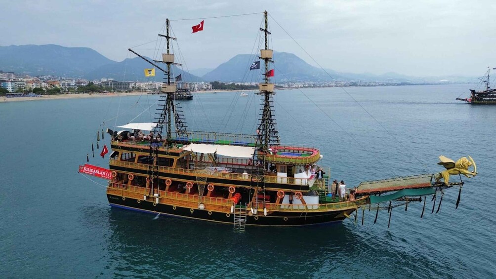 A large, multi-decked ship with multiple flags, docked at a harbor with mountains in the background. 4