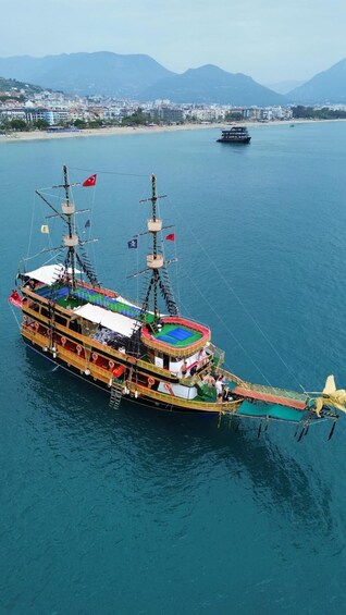 A large, multi-decked ship with multiple flags, sailing on a calm sea with a cityscape and mountains in the background. 3