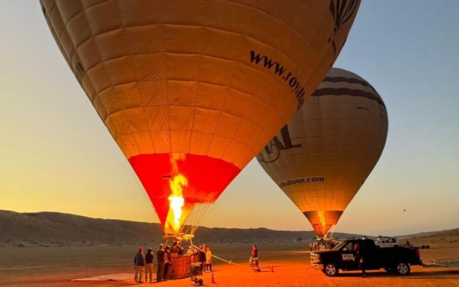 Two hot air balloons with flames at the base, a group of people, and a vehicle on a desert landscape. 1