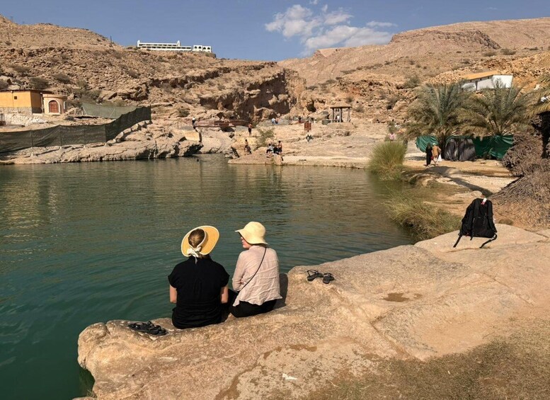 Two people sitting by a calm river with rocky terrain and a distant building. 5