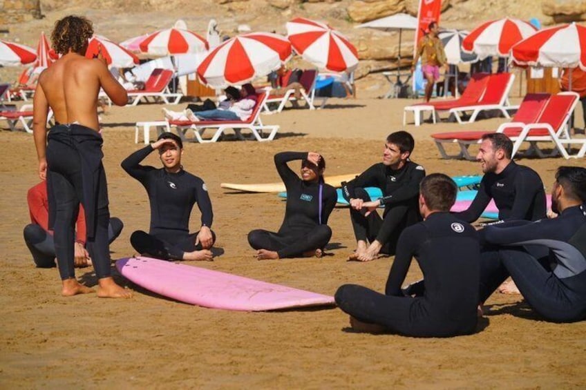 A group of people in wetsuits sitting on the beach, with surfboards and beach umbrellas in the background. 1
