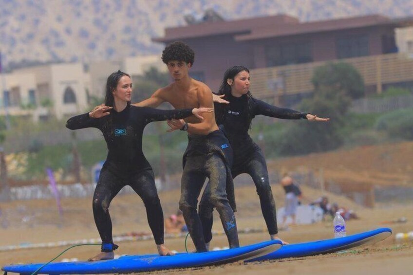 Drei Personen in Neoprenanzügen auf einem blauen Surfbrett an einem sandigen Strand, im Hintergrund sind Gebäude zu sehen. 1
