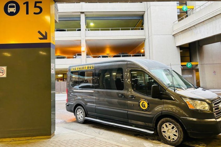 A gray van with 'The Fairy Bus' written on the side parked in front of a building with a yellow sign indicating a parking area. 2