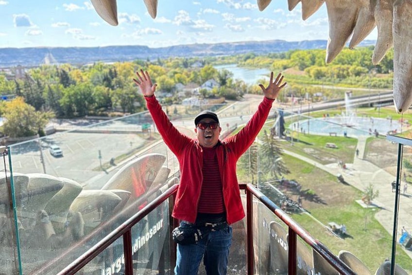 A person standing on a glass railing with arms raised, overlooking a landscape with trees, a body of water, and a bridge. 1