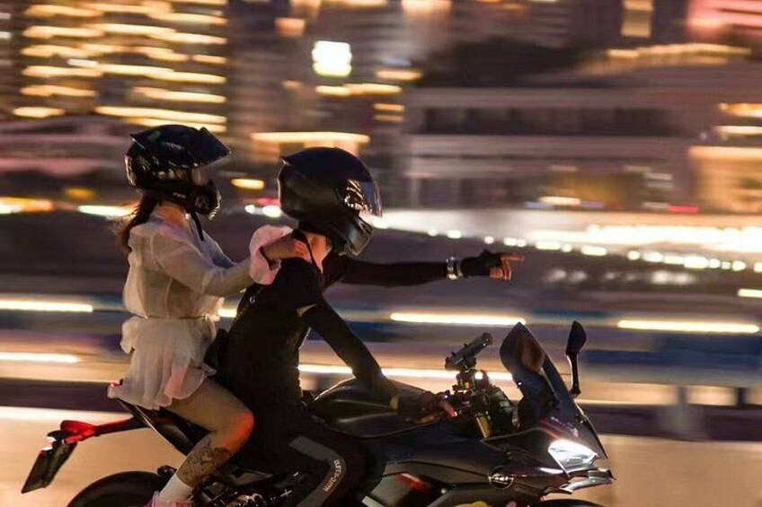 Two people on a motorcycle, one wearing a helmet, riding on a road with blurred lights in the background. 3