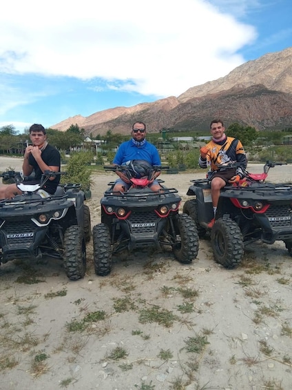 Trois personnes sur des quads dans une zone aride, avec des montagnes en arrière-plan. 2