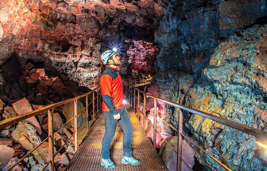 A person in a red jacket and helmet standing on a metal walkway inside a cave with colorful rock formations. 1