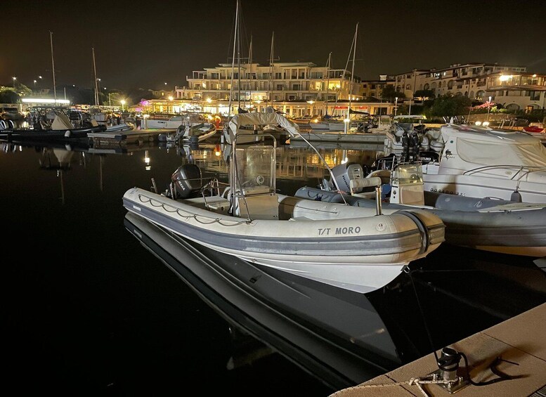 A marina at night with boats docked, a building with lights in the background, and calm water reflecting the scene. 2