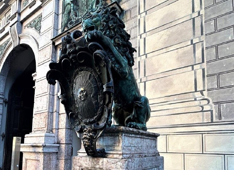 A bronze lion statue with a clock face on its chest, mounted on a stone pedestal, in front of a building with classical architecture. 2