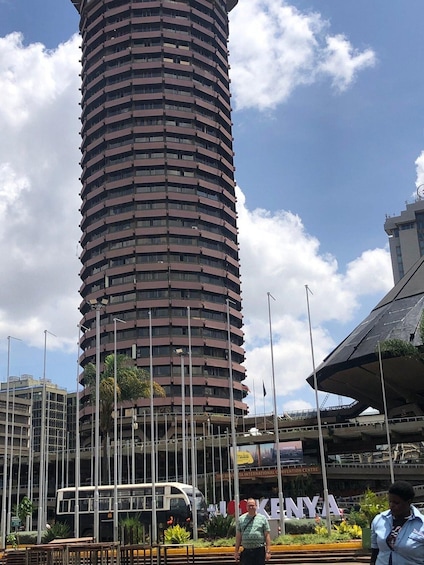 A tall cylindrical building with multiple balconies, a bus, and a sign for 'Kenya' in the foreground. 3