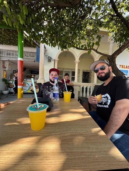 A group of people sitting at an outdoor table with drinks and food, under a tree with a building in the background. 3