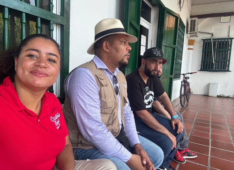 Three people sitting on a porch with green shutters and a bicycle in the background. 4
