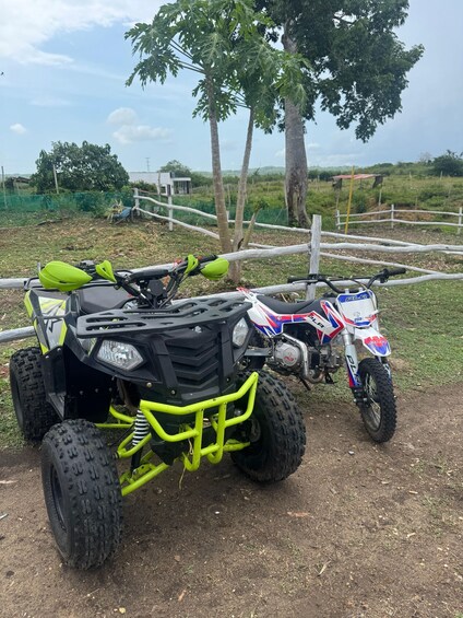 Two all-terrain vehicles parked on a dirt path with trees and a fence in the background. 2