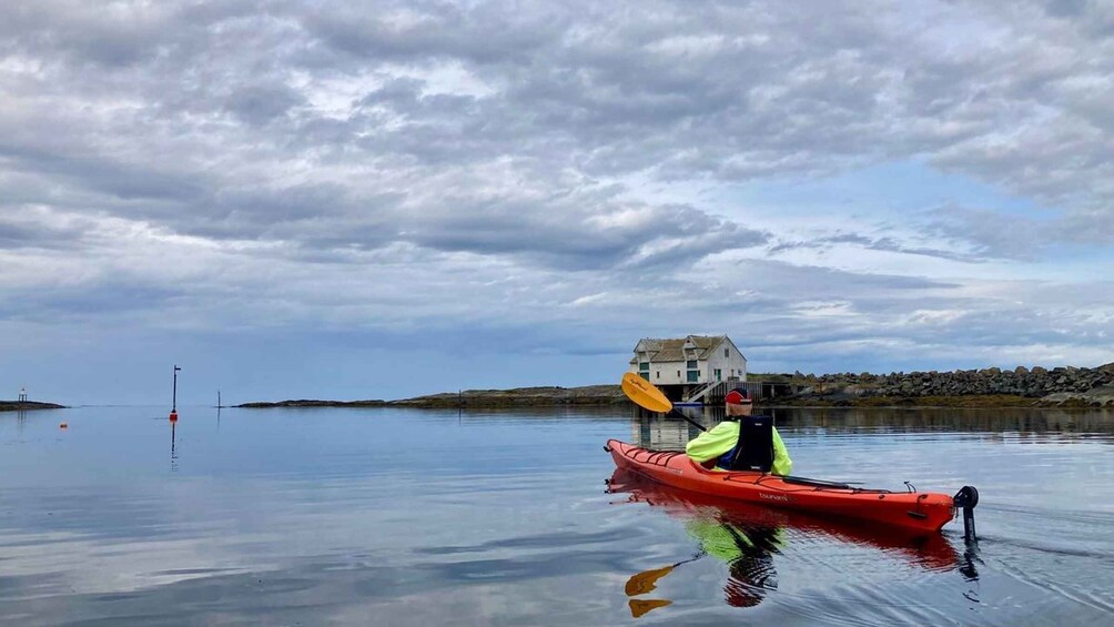 A person in a red kayak on calm water with a house and rocky shore in the background. 4