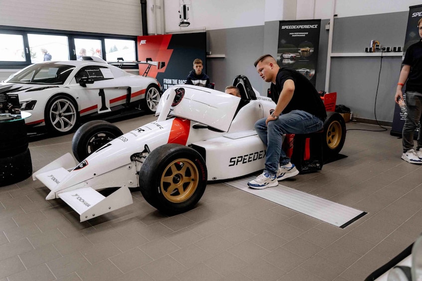 A person sitting on a racing car seat in a garage with a white race car and a red and white car in the background. 1