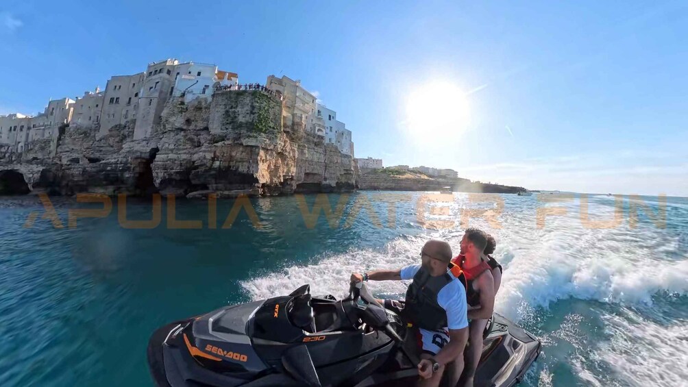 A jet ski on calm turquoise waters with a rocky coastline and white buildings in the background. 2