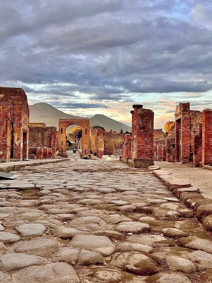 An ancient stone pathway leading through ruins with arches and brick structures, under a cloudy sky. 1