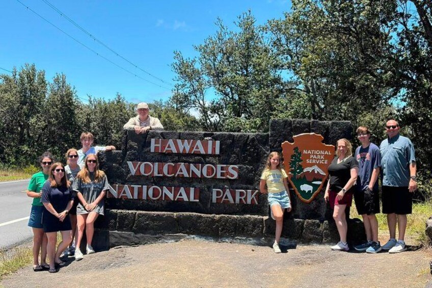 A group of people standing in front of a sign for Hawaii Volcanoes National Park. 1