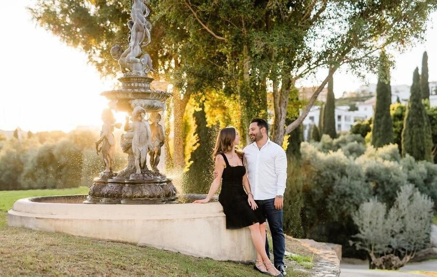 Ein Paar steht an einem Brunnen, im Hintergrund sind Statuen und Grünflächen zu sehen. 5