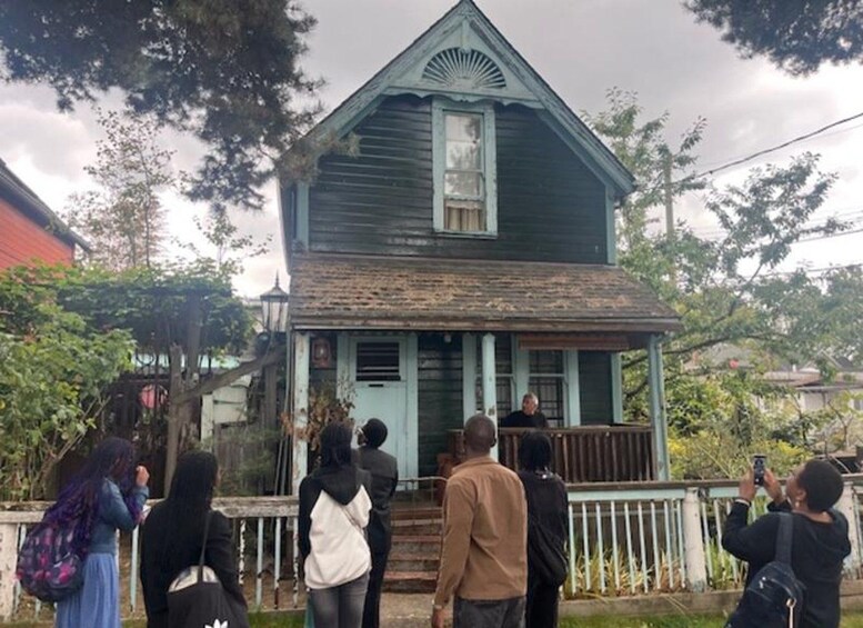 A group of people standing in front of a weathered, green wooden house with a porch. 2