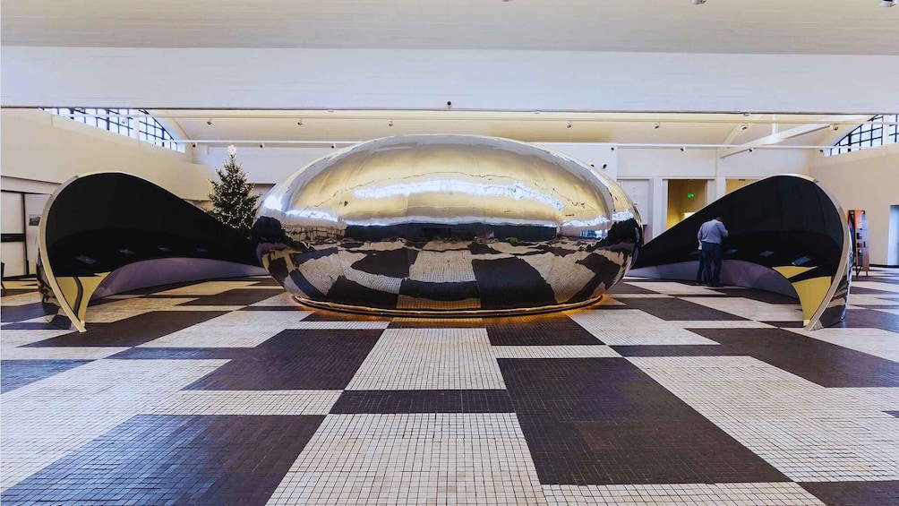 A large metallic sphere sculpture in a spacious indoor area with checkered flooring and two black and yellow seating areas. 1