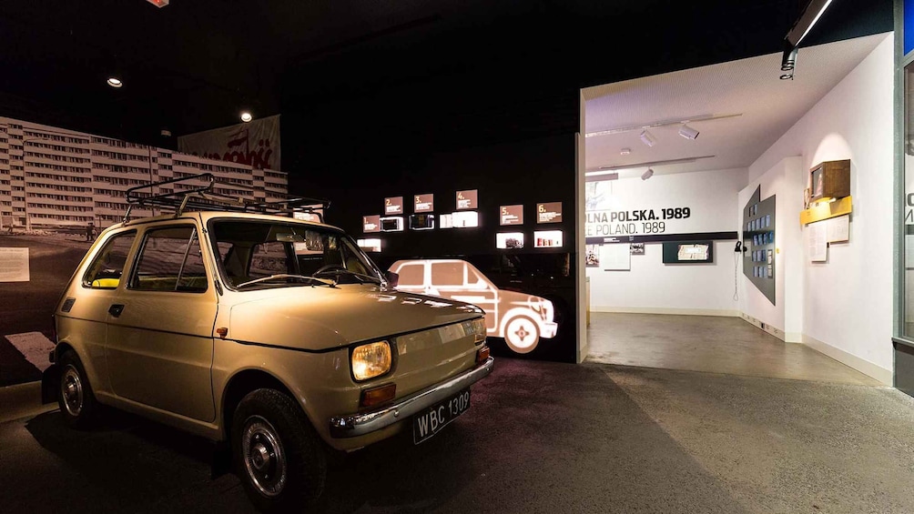 A vintage car on display with a backdrop of a building and informational panels. 5