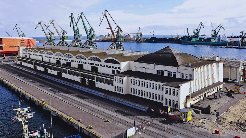 A large white building with a sloped roof, located near a port with multiple cranes and ships. 3