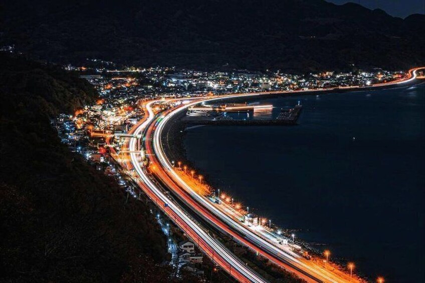 Una autopista costera de noche con estelas de luz dejadas por vehículos, un puerto deportivo y un pueblo a lo largo de la costa. 5