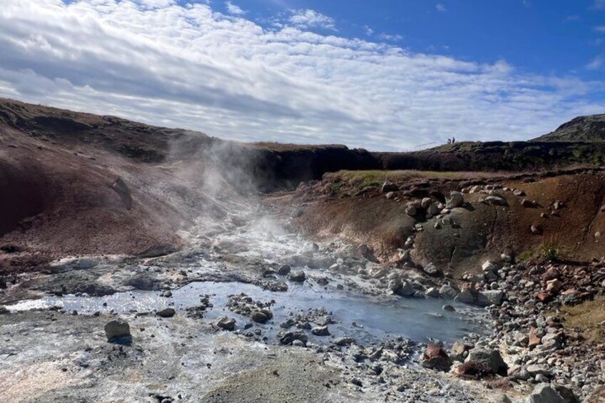 A geothermal area with steam rising from the ground, surrounded by rocky terrain and sparse vegetation. 2