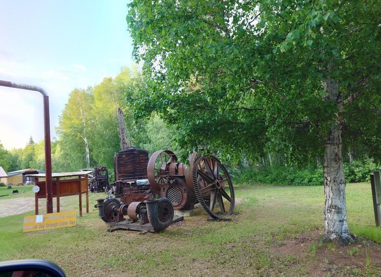 An outdoor exhibit of industrial machinery, including large gears and a crane, surrounded by trees and a grassy area. 2