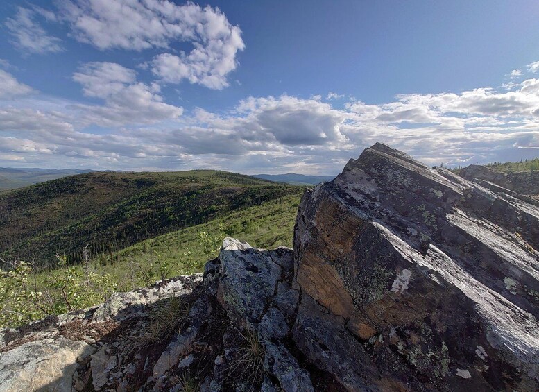 A mountainous landscape with a prominent rock formation in the foreground and a forested valley in the background under a partly cloudy sky. 1