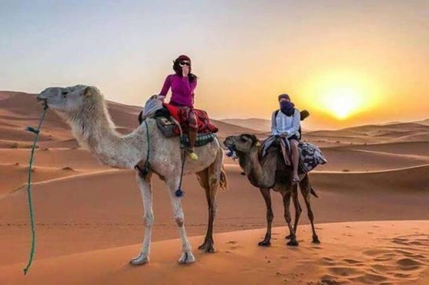 Two people riding camels in a desert landscape with sand dunes and a sunset in the background. 1