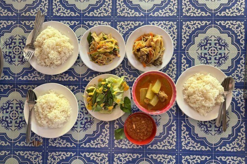 A table with six plates of food, including rice and various dishes, on a blue patterned tablecloth. 3