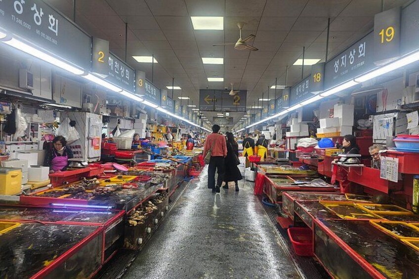 Un mercato con bancarelle che vendono diversi articoli, persone che passeggiano e cartelli sopraelevati che indicano i numeri delle bancarelle. 2