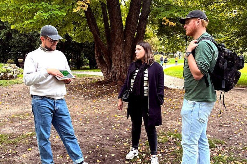 Tres personas de pie en un parque con árboles y suelo cubierto de hojas. 3