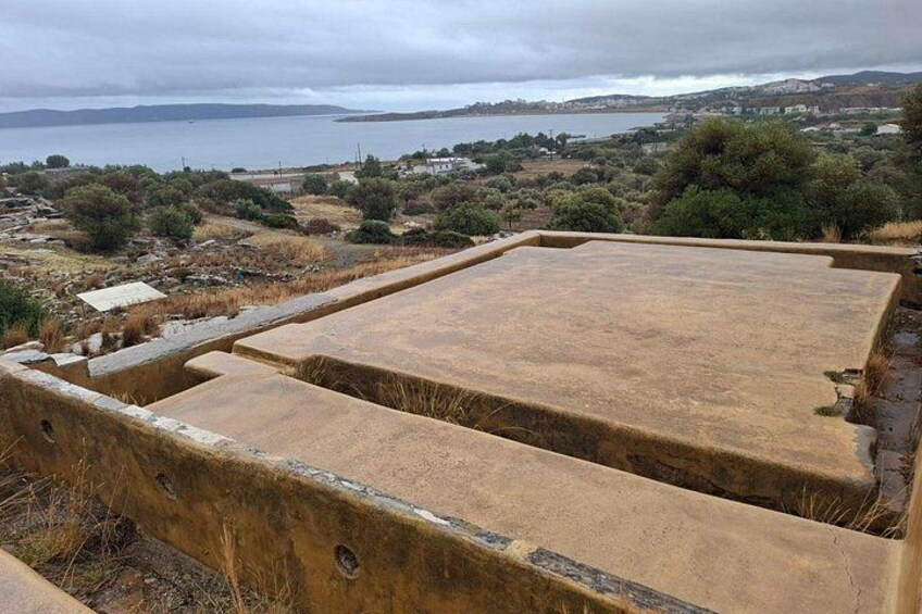 Un paysage côtier avec une grande structure en béton, peut-être un bâtiment ou un monument, dominant une étendue d’eau et des collines lointaines. 3
