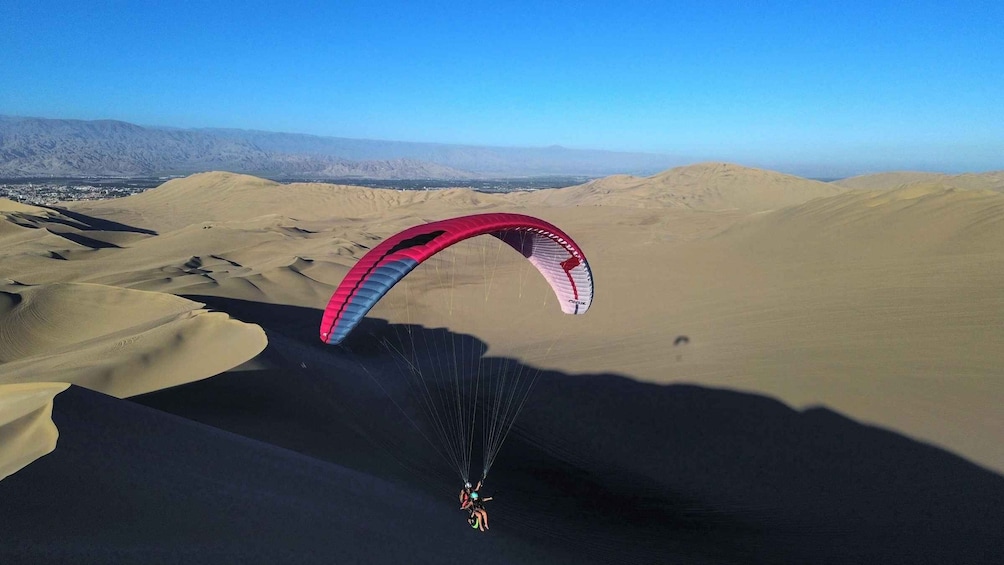 A person paragliding over a desert landscape with sand dunes and mountains in the background. 1