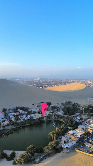 A coastal cityscape with a prominent pink kite flying over a body of water, surrounded by sand dunes and urban buildings. 3