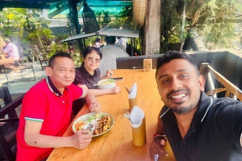 Three people sitting at an outdoor table with food and drinks, surrounded by greenery and other tables. 4
