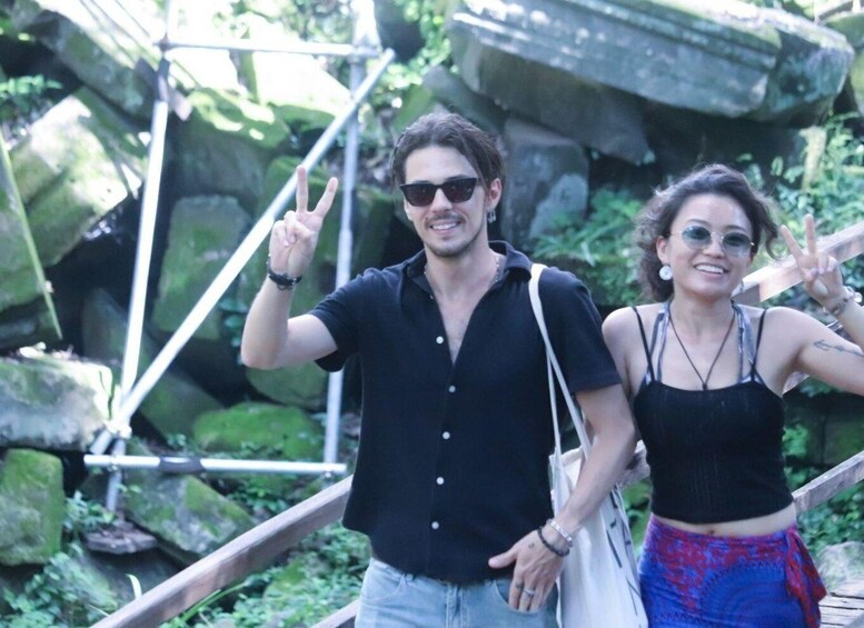 Two people walking on a wooden bridge with greenery and rocks in the background. 3