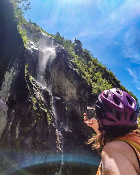 A person taking a photo of a waterfall with a helmet, surrounded by greenery. 5