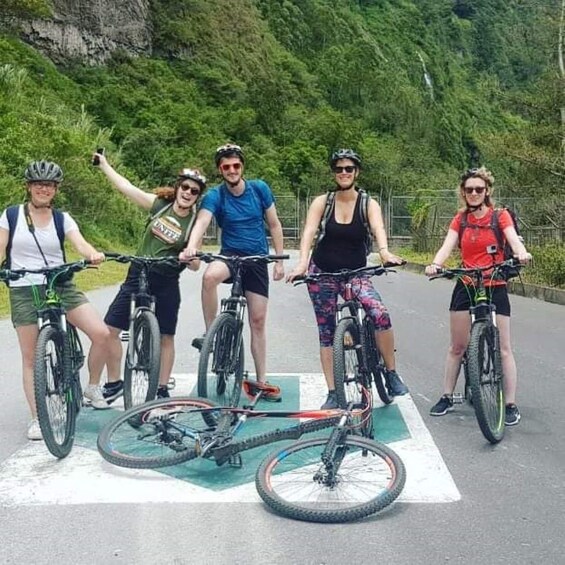 A group of cyclists on a paved path with a green bike lane, surrounded by lush greenery and a mountainous backdrop. 3