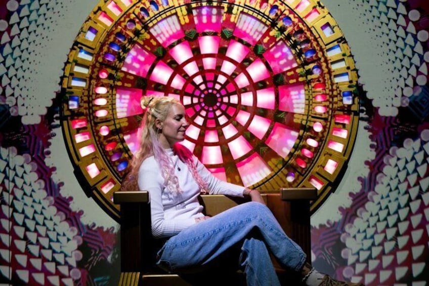 A person seated in front of a large, colorful circular structure with a patterned wall in the background. 4