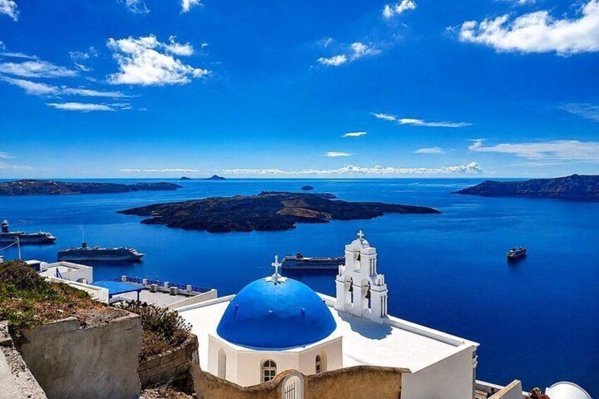 A coastal view with a prominent white building with a blue dome, a white bell tower, and a clear blue sea with boats. 1