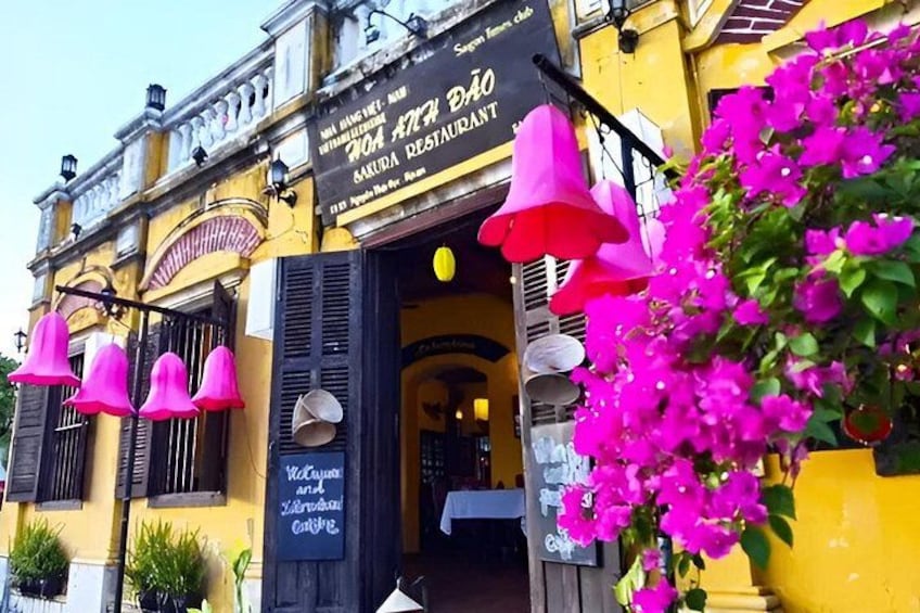 A yellow building with a sign for a restaurant, pink hanging decorations, and a pink flower bush. 5