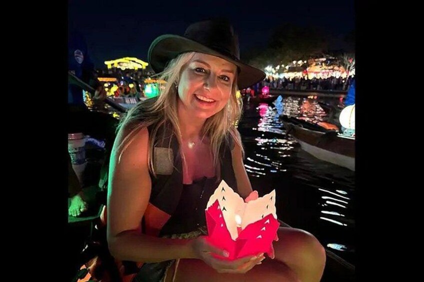 A person holding a red object with a light source inside, sitting on a boat with a water body and illuminated structures in the background. 1