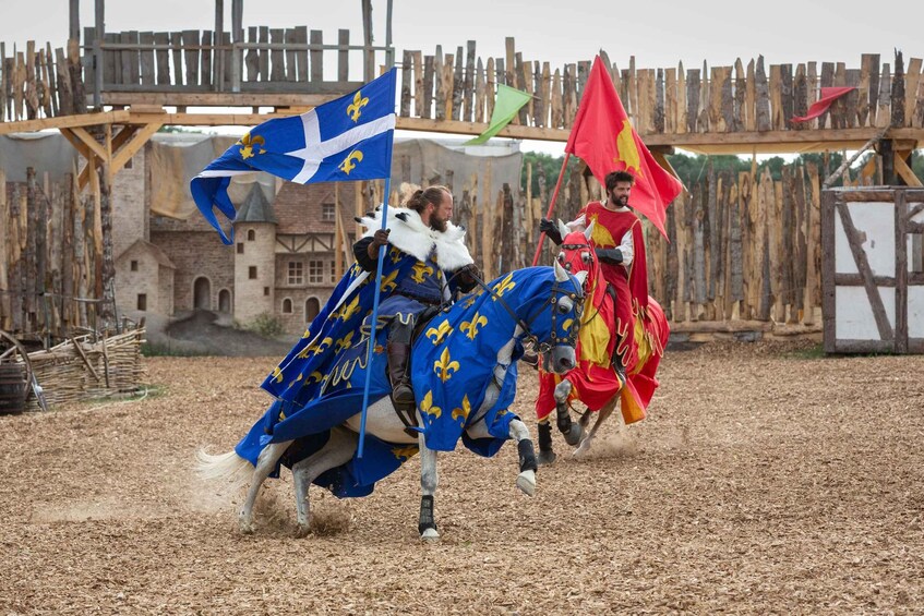 A medieval reenactment with participants on horseback, flags, and a wooden structure in the background. 1
