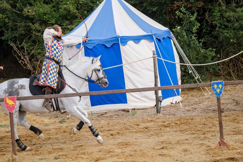 A knight in medieval costume riding a white horse with a checkered saddle, passing a blue and white tent. 2